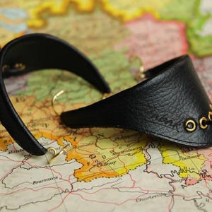 Puede incluir: Un par de pendientes de cuero negro con forma curva y semicircular. Cada pendiente presenta ojales de metal dorado y un fino lazo de cordón negro. La superficie de cuero negro texturizado es visible, mostrando el diseño de la pieza.