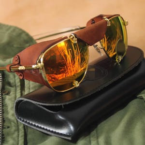 May include: Gold-rimmed sunglasses with reflective orange lenses and brown leather side shields. The sunglasses are resting on a black leather case. The lenses feature a metallic finish that reflects the surrounding environment in bright orange tones.