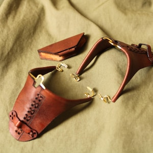 May include: Two brown leather hand guards featuring decorative lacing and brass-colored metal hooks, accompanied by a small, triangular brown leather case with a flap closure. The items are constructed from smooth, dark brown leather material.
