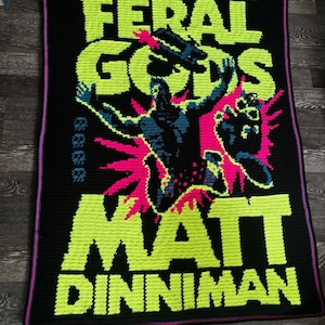 May include: A rectangular crochet blanket with a black background featuring neon green text that reads THE GATE OF THE FERAL GODS MATT DINNIMAN. A central figure in dark blue with bright pink accents is depicted in a dynamic pose against the text.