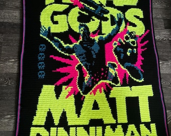May include: A rectangular crochet blanket with a black background featuring neon green text that reads THE GATE OF THE FERAL GODS MATT DINNIMAN. A central figure in dark blue with bright pink accents is depicted in a dynamic pose against the text.