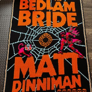 May include: A rectangular crochet blanket featuring a black background with orange text reading THE EYE OF THE BEDLAM BRIDE and MATT DINNIMAN. The design includes a light blue spiderweb, a central orange circle, a pink cat, and a pink figure.