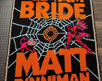 Può includere: Una coperta rettangolare all'uncinetto con sfondo nero e testo arancione che recita THE EYE OF THE BEDLAM BRIDE e MATT DINNIMAN. Il design include una ragnatela azzurra, un cerchio arancione centrale, un gatto rosa e una figura rosa.