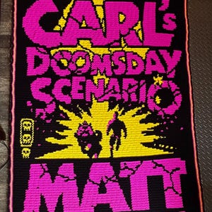May include: A rectangular crochet blanket featuring a black background with bright pink and yellow text and graphics. The text reads CARL'S DOOMSDAY SCENARIO, MATT, and DINNIMAN. The design includes a yellow sunburst and two silhouettes.