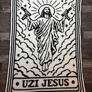 May include: A rectangular crochet blanket featuring a black and white graphic design of a figure holding two submachine guns. The central figure is surrounded by clouds, stars, and a halo. The text UZI JESUS is printed at the bottom.
