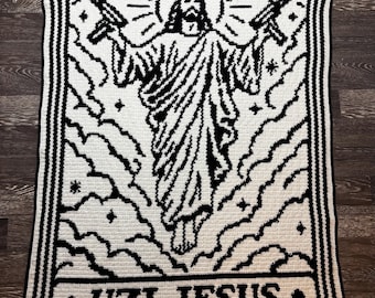 Può includere: Una coperta rettangolare all'uncinetto con un design grafico in bianco e nero di una figura che tiene in mano due mitra. La figura centrale è circondata da nuvole, stelle e un'aureola. Il testo UZI JESUS è riportato nella parte inferiore.