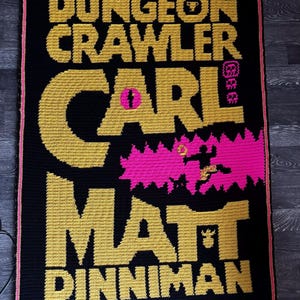 Puede incluir: Una manta de ganchillo rectangular con fondo negro y letras doradas que dicen DUNGEON CRAWLER CARL MATT DINNIMAN. El diseño presenta un elemento gráfico rosa brillante con una figura de silueta y un pequeño icono rosa cerca de la palabra CARL.