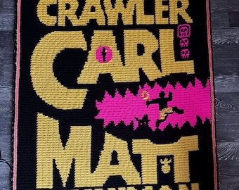 Può includere: Una coperta rettangolare all'uncinetto con sfondo nero e scritte dorate che recitano DUNGEON CRAWLER CARL MATT DINNIMAN. Il design presenta un elemento grafico rosa acceso con una figura sagomata e una piccola icona rosa vicino alla parola CARL.