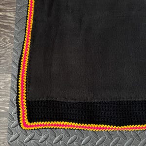 May include: A black fleece blanket featuring a wide black crocheted border along the edge, finished with a vibrant neon pink and bright yellow crocheted trim that frames the entire perimeter of the soft fabric material.
