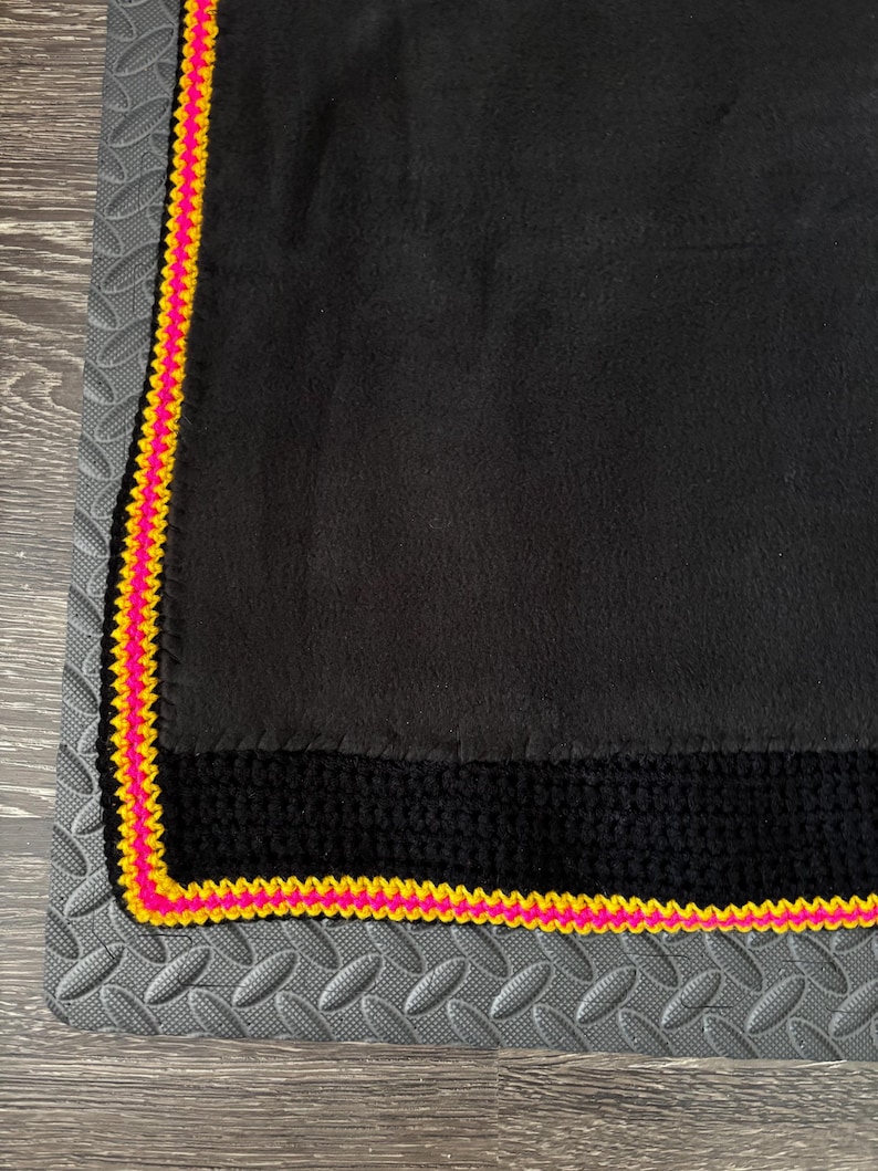 May include: A black fleece blanket featuring a wide black crocheted border along the edge, finished with a vibrant neon pink and bright yellow crocheted trim that frames the entire perimeter of the soft fabric material.