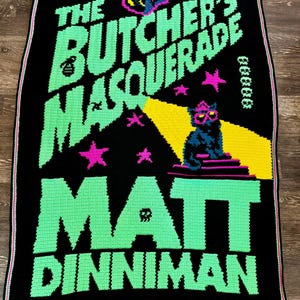 May include: A black rectangular crochet blanket featuring the text THE BUTCHER'S MASQUERADE and MATT DINNIMAN in large green letters. A black cat wearing pink glasses sits on a purple platform under a yellow spotlight, surrounded by pink stars.