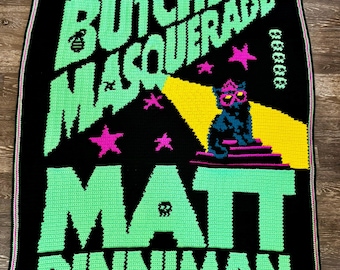 May include: A black rectangular crochet blanket featuring the text THE BUTCHER'S MASQUERADE and MATT DINNIMAN in large green letters. A black cat wearing pink glasses sits on a purple platform under a yellow spotlight, surrounded by pink stars.