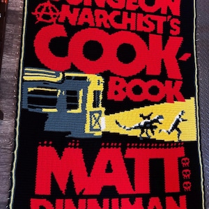 May include: A rectangular crochet blanket featuring a black background with red text that reads THE DUNGEON ANARCHIST'S COOKBOOK. It includes a yellow and grey graphic of a train with a headlight beam illuminating a small creature and a person, plus MATT DINNIMAN.