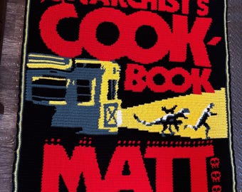 May include: A rectangular crochet blanket featuring a black background with red text that reads THE DUNGEON ANARCHIST'S COOKBOOK. It includes a yellow and grey graphic of a train with a headlight beam illuminating a small creature and a person, plus MATT DINNIMAN.