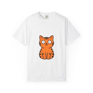 Peut inclure: Un t-shirt blanc à manches courtes arborant un motif central représentant un chat tigré orange avec des rayures noires, de grands yeux blancs et un petit sourire. Le chat est dessiné dans un style cartoon avec un contour noir épais.