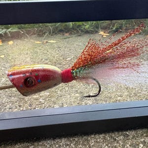 May include: A fishing lure featuring a rounded, glittery gold and red head with a dark eye and small blue accents. A thin wooden stick protrudes from the front. The back consists of long, shimmering red and white synthetic fibers tied with red thread.
