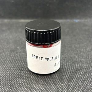 Op de afbeelding: Een kleine, doorzichtige cilindrische pot met een zwart geribbeld schroefdeksel. De pot bevat een rode substantie die door het glas zichtbaar is. Een wit etiket op de voorzijde bevat de zwarte gedrukte tekst BOOTY HOLE RED en B'NG in een schreefloos lettertype.