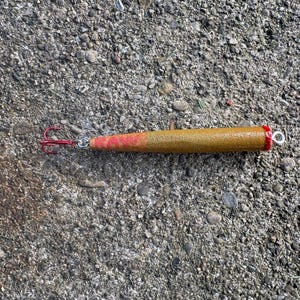 Puede incluir: Un señuelo de pesca cilíndrico con un cuerpo texturizado de color dorado-marrón que presenta la palabra RAW en letras rojas. El señuelo tiene un anzuelo triple rojo unido a un extremo con un eslabón giratorio y un pequeño ojal plateado.