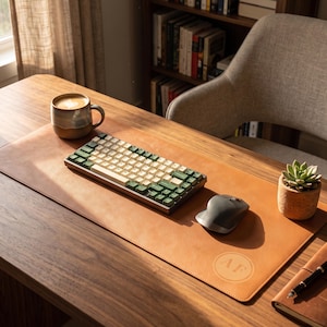 Könnte beinhalten: Eine beigefarbene Schreibtischunterlage aus Leder mit einem kreisförmigen geprägten Logo mit den Buchstaben AF, einer mechanischen Tastatur mit cremefarbenen und dunkelgrünen Tasten, einer grauen Funkmaus, einer Keramiktasse mit Kaffee und einer kleinen Sukkulente.