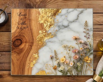 Op de afbeelding: Een rechthoekige serveerplank met een gespleten ontwerp van natuurlijk houtnerf en wit marmer, verbonden door een bladgouden accent. De houten kant is gegraveerd met de tekst Lucas & Mia 12.20.2026. De marmeren kant toont geperste wilde bloemen.