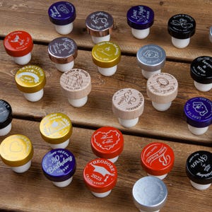 May include: A collection of round bottle stoppers with white silicone bases and colorful metal or wooden tops. Each top features unique laser-engraved designs, including names, dates, graduation caps, hearts, rings, and animal illustrations.