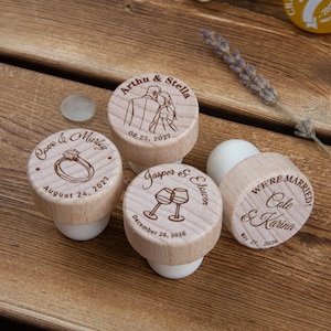May include: Four round wooden bottle stoppers with white silicone bases, each featuring custom laser-engraved designs including names, dates, and illustrations of a couple, a ring, wine glasses, and the text WE'RE MARRIED, Cole & Karina.