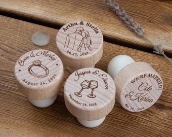 Könnte beinhalten: Vier runde Weinflaschenverschlüsse aus Holz mit weißen Silikonunterteilen, jeweils mit individuellen Lasergravuren von Namen, Daten und Illustrationen eines Paares, eines Rings, Weingläsern sowie dem Text WE'RE MARRIED, Cole & Karina.