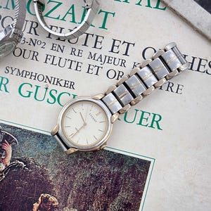 Puede incluir: Un reloj de pulsera bicolor con caja redonda plateada, bisel dorado y esfera color crema con la palabra "LUCENT" en texto negro. El reloj cuenta con una correa de eslabones metálicos que alterna segmentos de color plateado y dorado.