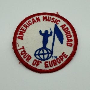 May include: A circular embroidered patch with a white background and a red border. The text AMERICAN MUSIC ABROAD TOUR OF EUROPE is stitched in red. A blue graphic shows a figure standing on a globe, holding a baton next to a flag.