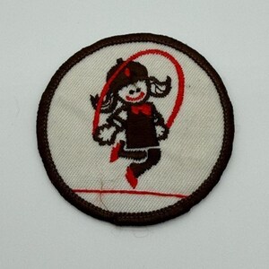 May include: A circular embroidered patch with a white background and a dark brown border. The design features a stylized figure in dark brown clothing with red accents, jumping rope with a red rope. The figure has a simple face and hair.