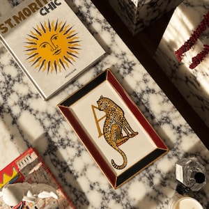 Könnte beinhalten: Ein rechteckiges Keramiktablett mit einer zentralen Illustration eines Leoparden, der neben einem großen goldenen Buchstaben A sitzt. Das Tablett hat eine weiße Mitte, einen dunkelroten Rand und schwarze Ecken mit einer goldenen Verzierung.