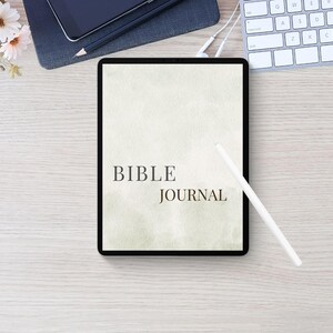 May include: A digital tablet displaying the text BIBLE JOURNAL in dark serif font on a pale, textured, light green background. A white stylus rests diagonally across the screen of the tablet, which is positioned on a light wood surface.