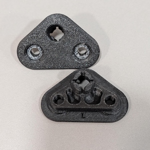 May include: Two black, triangular, 3D-printed plastic components with textured surfaces. Each piece features a central circular opening with notched edges and two smaller circular holes. One piece is marked with the letter L on its base.