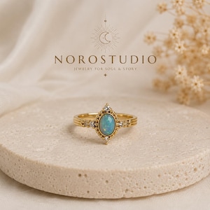 May include: A gold ring featuring a central oval-shaped iridescent opal stone surrounded by small clear stones in a beaded setting. The text NOROSTUDIO JEWELRY FOR SOUL & STORY is visible above the ring, along with a crescent moon icon.