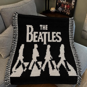 May include: A black and white crocheted blanket featuring the text THE BEATLES in a bold font above a graphic silhouette of four figures walking in a line. The blanket has a textured stitch pattern and is finished with a black and white fringe.