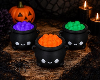 May include: Three black cauldron-shaped containers with smiling faces, each filled with a textured substance in bright purple, orange, or green. The containers feature small side handles and rounded bases, presenting a cheerful appearance.