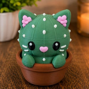 May include: A green, 3D-printed cat-shaped cactus figurine with white prickle details, pink heart-shaped nose, and pink inner ears. The cat figure has large black eyes and white whiskers, and it is seated inside a small, round, brown terracotta-colored pot.