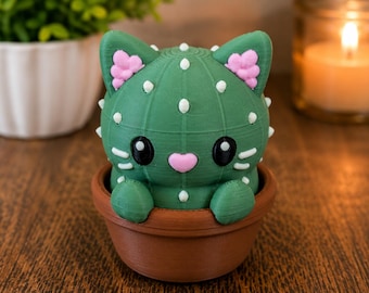 May include: A green, 3D-printed cat-shaped cactus figurine with white prickle details, pink heart-shaped nose, and pink inner ears. The cat figure has large black eyes and white whiskers, and it is seated inside a small, round, brown terracotta-colored pot.