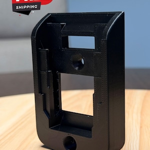 May include: A black, 3D-printed rectangular mounting bracket featuring a hollow center, a circular opening at the bottom, and a central circular hole. The structure has a textured surface with visible layer lines and a flat, open-faced design.