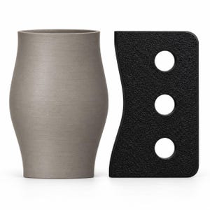 May include: A matte grey cylindrical vessel with a slightly curved profile positioned next to a rectangular black tool featuring a textured surface and three circular cutouts arranged in a vertical line along its length.