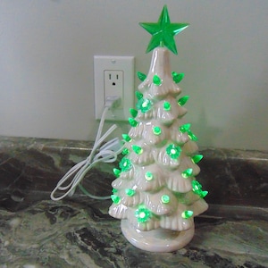 May include: A ceramic Christmas tree with green lights and a green star on top. The tree is off-white and has a glossy finish. The lights are shaped like flowers. The tree is plugged into an electrical outlet.