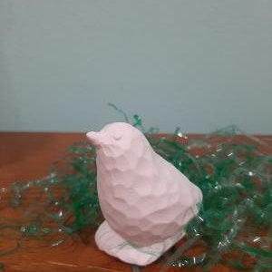 May include: A small, white, ceramic bird figurine with a rounded body and a slightly upturned beak. The bird is sitting on a bed of green plastic grass.