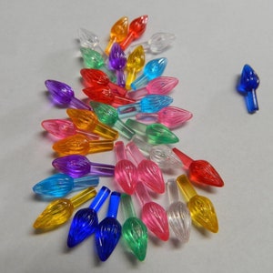 May include: A collection of colorful, transparent Christmas light bulbs in various colors including red, orange, yellow, green, blue, pink, and purple. The bulbs are arranged on a light gray surface.