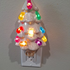 May include: A white ceramic Christmas tree night light with colorful lights. The tree is plugged into an electrical outlet.