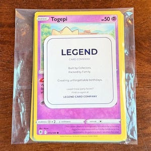 Puede incluir: Una carta coleccionable de Togepi con 50 HP, dentro de una funda de pl&aacute;stico transparente. Un inserto rectangular blanco est&aacute; colocado sobre la carta, con el texto LEGEND CARD COMPANY, Built by Collectors, Packed by Family y Creating unforgettable birthdays.