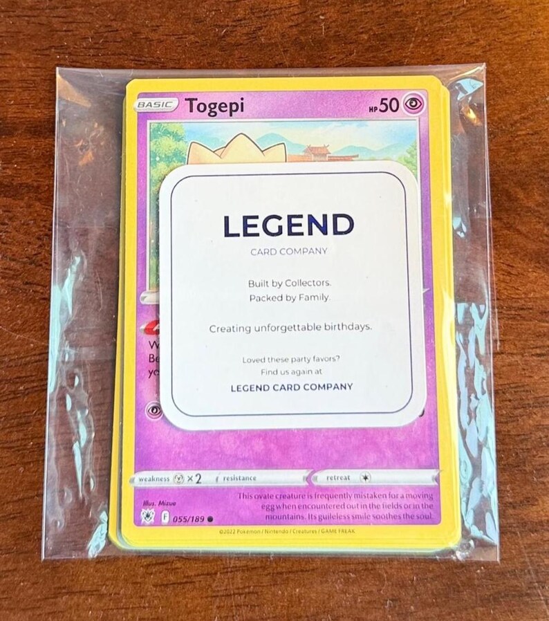 Puede incluir: Una carta coleccionable de Togepi con 50 HP, dentro de una funda de pl&aacute;stico transparente. Un inserto rectangular blanco est&aacute; colocado sobre la carta, con el texto LEGEND CARD COMPANY, Built by Collectors, Packed by Family y Creating unforgettable birthdays.