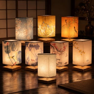Peut inclure: Une collection de huit lampes de table rectangulaires avec des bases en bois et des abat-jours en tissu. Chaque abat-jour présente des illustrations imprimées distinctes, notamment des cerisiers, des lotus, des oiseaux et des paysages.