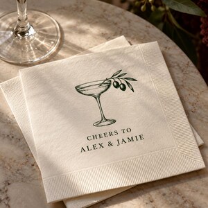Ivory paper cocktail napkins printed with a dark green coupe-glass and olive-sprig illustration and sample wording Cheers to Alex & Jamie. The folded paper edges are visible beside a glass. Digital design mockup.