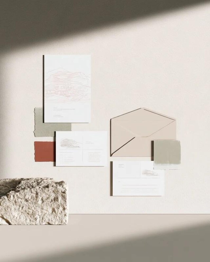 Coordinated wedding invitation stationery with cream cards, muted green accents and an envelope, arranged on a pale background. Fine architectural illustration and small printed text appear on the cards. Digital concept mockup.