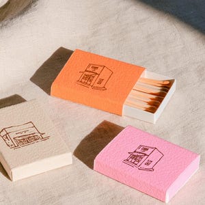 以下が含まれることがあります： オレンジ、ピンク、クリーム色の3つの長方形のマッチ箱。それぞれに店舗の茶色の線画イラストが描かれています。オレンジ色の箱は半分開いており、白い内箱の中に茶色の頭を持つ木製のマッチが見えています。
